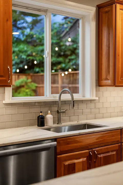 Kitchen window over the sink looking out to the lit backyard at dusk